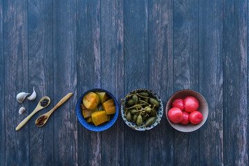 Assorted pickles: aubergines, spring onions and capers in bowls on a wooden surface, top view, copy space.