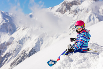 Fototapeta na wymiar Beautiful girl in pink helmet and ski equipment sit on top of the mountain in snow observing the view