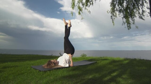 Young fit woman practice yoga on coast near the lake or sea. Woman doing Supported Shoulder Stand Pose