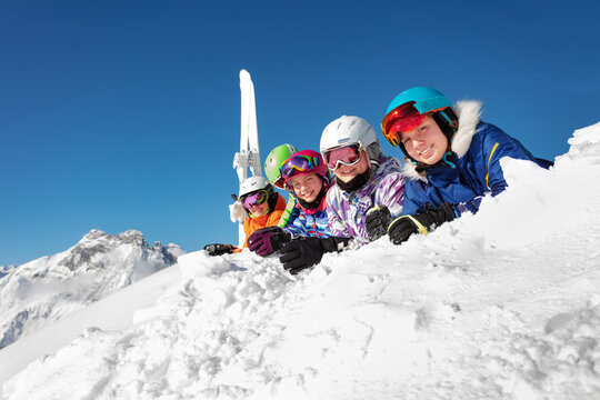 Big group of ski kids lay on snow cheerfully smiling over mountain peak and blue sky in colorful sport outfit