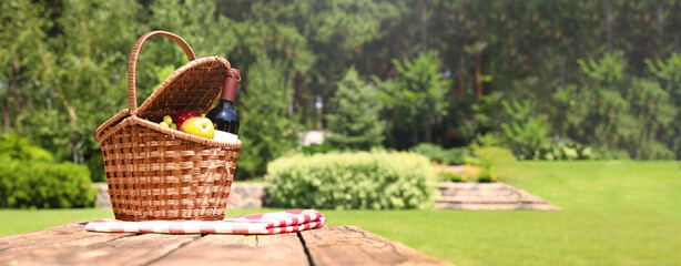 Picnic basket with fruits, bottle of wine and checkered blanket on wooden table in garden, space...