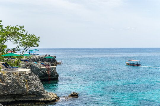 Tour boat sailing by Rockhouse Hotel on the cliffs of Negril, Westmoreland in Jamaica. Famous vacation spot with seaside scenic views on the island coast. Outdoor lounge chairs & umbrellas