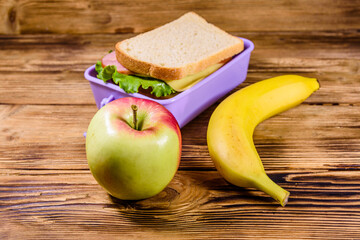 Ripe apple, banana and lunch box with sandwiches on a wooden table