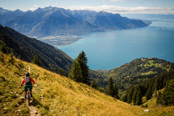 Fototapeta na wymiar MONTREUX, SWITZERLAND. A woman rides a mountain bike along a narrow trail across a steep grassy Alpine pasture above Montreux and Lake Geneva.