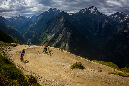 LES DEUX ALPES, FRANCE. Two mountain bikers riding a 'bermed' corner on a bike-park trail, with a steep river valley and dramatic alpine peaks behind.