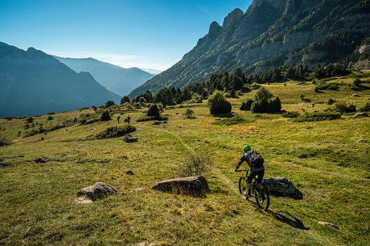 BIESCAS, HUESCA, SPAIN. A group of mountain bikers riding a narrow trail through open grassland with dramatic peaks and blue sky behind.