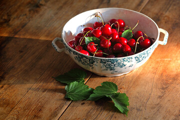 beautiful white and blue ceramic bowl filled with juicy scarlet red cherries on a dark old wood kitchen buffet of old traditional cuisine, green leaves and a ray of warm light