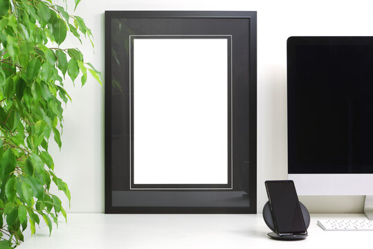 Empty frame to personalize placed on a white desk next to a computer, smartphone and a potted ficus tree, for staging a photo or drawing