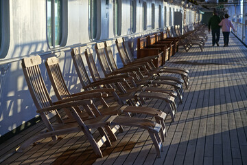 An elderly couple strolls past a long row of empty deck chairs on the Promenade Deck of a cruise ship.