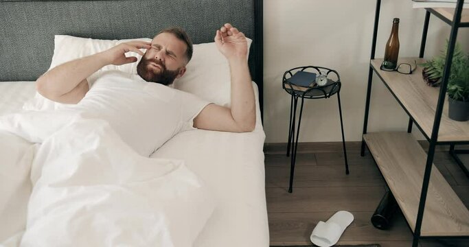 Tired bearded young man waking up in bed and having hangover with head pain. Handsome adult guy in 30s grimacing and touching head with both hands. Concept of bad morning.
