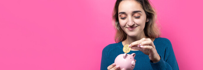 Girl puts in the piggy bank physical Bitcoin.Young girl over pink background holding piggy bank and rejoices gesturing. The concept of reliability of cash investments and insurance.