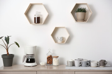 Modern coffeemaker and cups on commode near light wall indoors