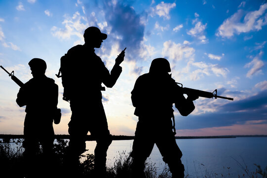 Silhouettes of soldiers with assault rifles and portable radio transmitter patrolling outdoors. Military service
