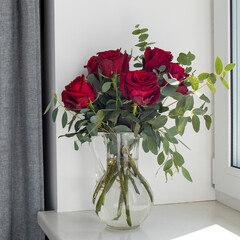 A wedding bouquet of nine roses and various varieties of eucalyptus in a glass jug on the windowsill.
