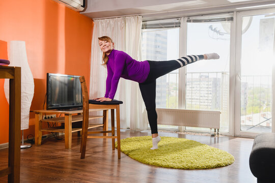 Elderly Smiling Woman Workout Exercise Stretching At Home Interior, Put Hands On Chair And Raise Leg Up, Wearing Sport Leggings, Isolation Gym Indoors