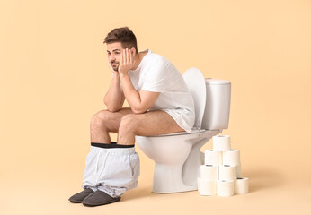 Young man with rolls of paper sitting on toilet bowl against color background