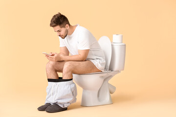 Young man with mobile phone sitting on toilet bowl against color background