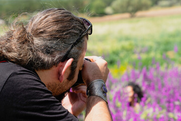 Long haired male photographer takes a photo of a girl in purple flowers