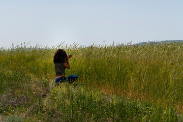 Naklejka na ściany i meble Back view of girl with black curly hair is sitting and taking a photo near a green and yellowish wheat field
