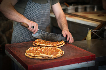 Baked lahmacun is a round, thin piece of dough topped with minced meat (most commonly beef or lamb), minced vegetables and herbs. Man cuts lahmacun.