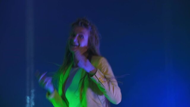 The woman dances merrily and looks at the camera in the light of strobe lights and spotlights. Neon colors and crazy girl dancing to music and singing