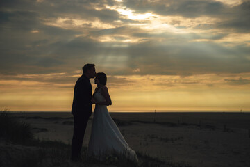 Silhouette of newlyweds on evening beach. Groom kissing bride on forehead. Dramatic sky with sun rays among clouds in background. Happiness, love