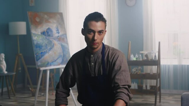Portrait of young confident man, artist in working apron, sitting in his beautiful bright studio, have just finished his work on canvas - a contrast landscape, inspired by visual art, Slow motion.