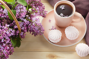 Cup of tea or coffee with sweet pink marshmallows and bouquet of lilac in a basket on wooden background. Breakfast in the morning.