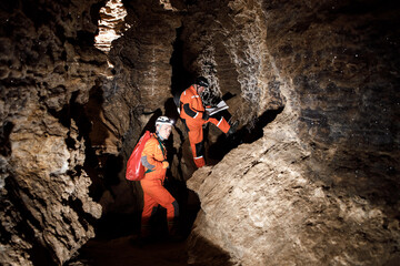 Two men, strong physique, explore the cave. Men dressed in special clothes to pass through the cave and stopped, looking at the map - obrazy, fototapety, plakaty