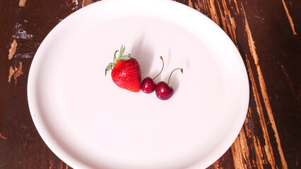 Top view of red strawberry and two sweet red cherries in the white plate on a wooden background. Space for text. Design concept.