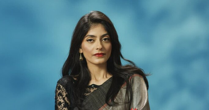 Portrait shot of Hindu young attractive serious offended and selfish woman in traditional clothes smiling to camera on blue wall background. Pretty stylish vain female in Indian style.