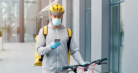 Deliveryman in medical mask, gloves and casque leading his bicycle at street, tapping on cellphone, courier having phone call.