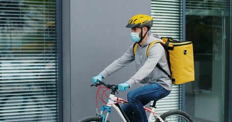 Young courier in medical mask delivering food with yellow thermal backpack, riding a bicycle in the city. Food delivery service concept