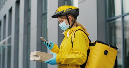 Portrait shot of beautiful Caucasian woman delivery worker in casque, gloves and medical mask standing at street and holding carton boxes. Pretty female courier with parcels looking at camera.