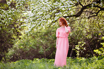 Young woman in romantic long pink dress in a blooming apple garden. Wind is hairs. Spring story. Red-haired girl smiles and plays with her hairs. 