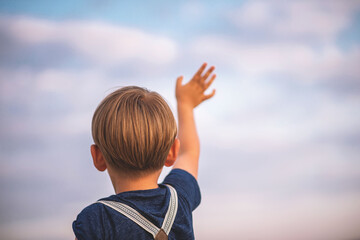 little boy shows his hand in a beautiful sky pink-purple sky in a blue T-shirt and suspenders rear view