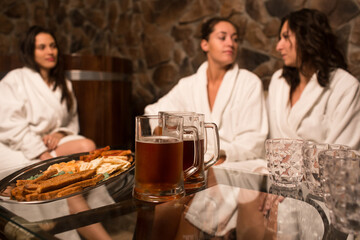 happy young caucasian women in bathrobe sit next to pool at sauna, drinking cold beer. 