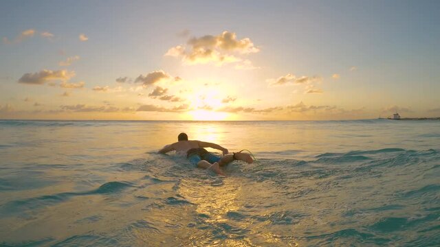 SLOW MOTION, CLOSE UP: Athletic man on surfing vacation in Barbados paddles out to the open sea to surf the last wave of the day. Cinematic shot of a male surfer paddling out toward the golden sunset.