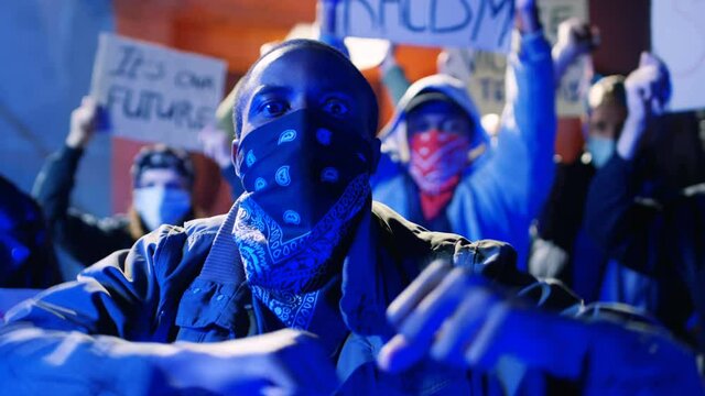 Portrait of young African American male protester with masked face screaming mottos and shouting slogans. Guy protesting at night in crowd against racism. Man rebel at riot against police aggression.