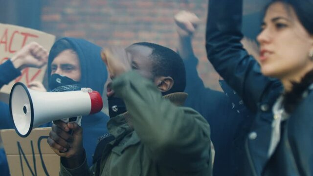 Male and female mixed-races pretesters screaming slogans and protesting in USA against racism and police acts. African American guy leading and shouting in megaphone and Caucasian girl holding poster.