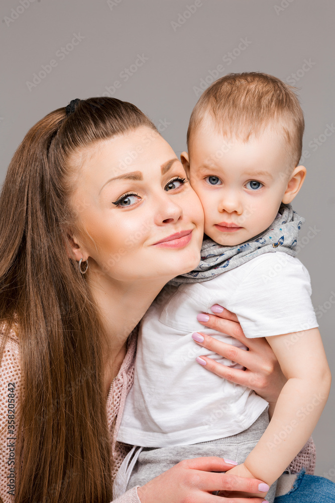 Wall mural Beautiful young mother and little sweet son embracing, woman gesturing and pointing at camera by finger. Smiling stylish family of mom and kid with blue eyes, posing at gray studio. Gray background.