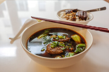 Selected focus view at Taiwanese stew Beef Noodle Soup without noodle in white bowl on white table and blur background of Slow cook Soy Sauce Braised Pork Belly, with rice.