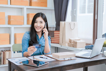 Asian beautiful businesswoman working from home in work office room, holding credit card concept of online payment transaction purchasing from ecommerce shopping online store, using computer laptop