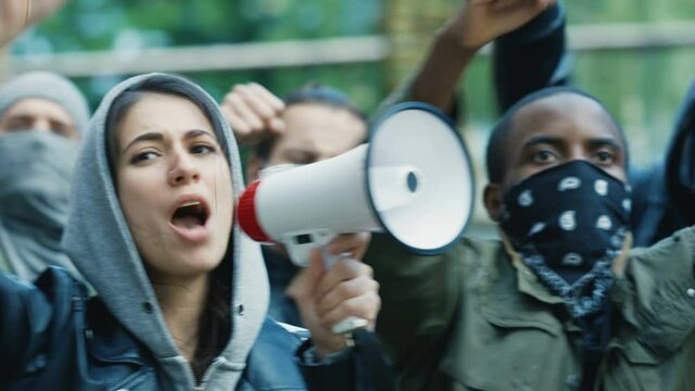 Caucasian young woman leader screaming mottos in megaphone at street in crowd and smoke at protest against racism in USA. Active mixed-races protesters shouting demands. Female leading at rebellion.