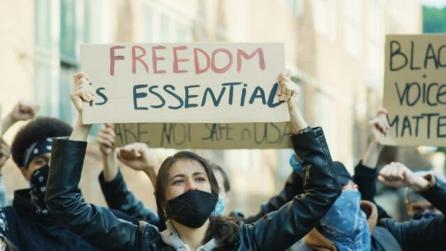 Beautiful Caucasian young woman in mask hoding poster Freedom essential and screaming mottos at protest in multiethnic crowd. Female protester at manifestation for human rights and against violence.
