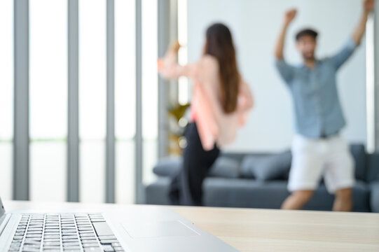 Laptop Notebook Computer On The Table With Happy And Relax Young Couple Lover Wearing Casual Dress Together Dancing In Living Room At Home.