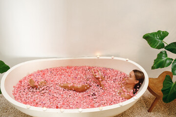 Dreamy young  woman lying in bath full of pink roses, hold in hands flower petals. Woman relaxing in round outdoor bath with tropical flowers. Organic skin care in luxury spa resort