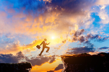 At sunrise, a male businessman silhouette leaps from one side of the cliff to the other.
