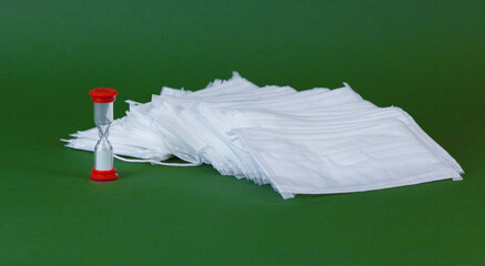 A stack of disposable medical masks and an hourglass on a green background, close-up.