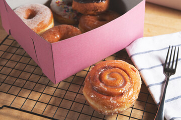 A view of a cinnamon roll next to a pink box full of donuts, in a still life setting.